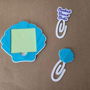 May include: A bright blue seashell-shaped sticky note holder containing a square light green sticky note, accompanied by two white paper clips, one featuring the text Bookish Beach Babe and the other topped with a small blue seashell.