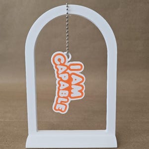 May include: A white arched display stand holds a hanging tag suspended by a silver ball chain. The tag is white with a thick orange border and features the orange text I AM CAPABLE in a bold, rounded, sans-serif font, oriented vertically.