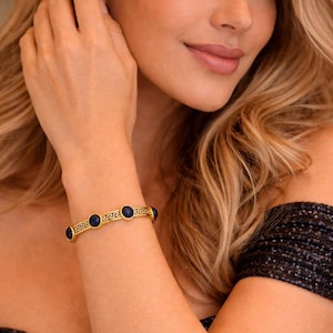 Puede incluir: Brazalete dorado con diseño de llave griega y cinco piedras preciosas redondas de color azul oscuro. El brazalete se lleva en la muñeca, con un top negro con lentejuelas en el fondo. Joyería.