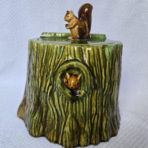 May include: A ceramic cookie jar shaped like a green tree stump with a textured bark finish. A brown squirrel figurine sits on the lid, and a second squirrel head is visible peeking out from a hollow opening on the front of the tree stump.