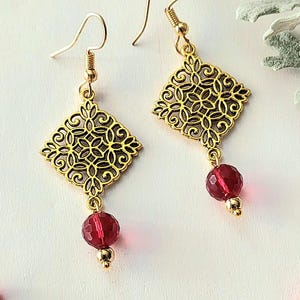 May include: Gold-coloured filigree earrings with a diamond-shaped design. Each earring features a faceted, red bead and a small gold bead. The earrings have a hook closure.