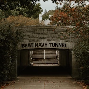 Puede incluir: Un túnel de piedra arqueado con las palabras "BEAT NAVY TUNNEL" sobre la entrada. El túnel conduce a una escalera. Los árboles con hojas verdes y rojas enmarcan la entrada, y un camino de hormigón conduce a ella.