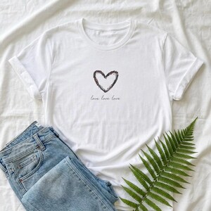 Puede incluir: Camiseta blanca con un diseño en forma de corazón y las palabras "love love love" impresas debajo. Se muestra con vaqueros azules y una hoja de helecho verde sobre un fondo blanco.