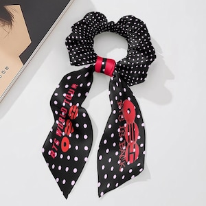 May include: Black scrunchie with white polka dots and red accents. The scrunchie has two long, black, polka-dotted tails with the text "BTS WORLD TOUR" and "BTS IS BACK" in red. The scrunchie is made of a silky material.