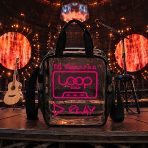 Könnte beinhalten: Eine durchsichtige Kunststofftasche mit schwarzem Rand und Tragegriffen, die mit dem leuchtend pinken Text Ed Sheeran, Loop Tour und dem Wort PLAY sowie einem Dreieckssymbol auf der Vorderseite bedruckt ist. Die Tasche hat seitliche Netztaschen.