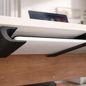 Pode incluir: Um suporte preto para tablet sob a mesa está montado na parte inferior de uma mesa branca. O suporte, feito de um material espesso e curvo, suporta um tablet branco. A mesa tem uma base de madeira.