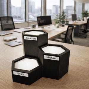Pode incluir: Um organizador de secretária preto com três compartimentos hexagonais, cada um com um interior branco. O organizador tem etiquetas com os nomes "Emily", "Jonathan" e "Michael". O organizador está numa secretária de madeira clara num ambiente de escritório.
