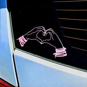 May include: Pink vinyl decal of two hands forming a heart shape. The hands have bracelets on the wrists. The decal is on a black background, likely a car window. The car is light blue.