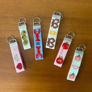 May include: Six rectangular keychains featuring cross-stitch embroidery designs. The patterns include green olives, red lobsters on blue, the letter B with a pretzel, red tomatoes on pink, and peach slices on light green, all with silver hardware.