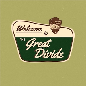 May include: A vintage-style sign featuring the words "Welcome to The Great Divide". The sign is predominantly green and white, with a brown emblem shaped like an arrowhead at the top. The background is a muted green.