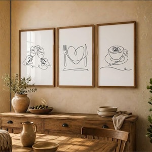 May include: Three framed wall art prints featuring black continuous line drawings on white backgrounds. The left print shows two clinking wine glasses, the center print displays a fork and knife forming a heart, and the right print depicts a coffee cup.