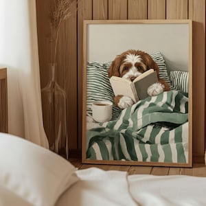 以下が含まれることがあります： ベッドで本を読む茶色と白の犬が描かれた額入りのプリント。犬は緑と白のストライプの毛布と枕の下にいます。白いマグカップが犬の隣のベッドに置かれています。