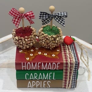 May include: A decorative stack of three wooden books painted red, green, and brown with the text "HOMEMADE CARAMEL APPLES" in white. Two faux caramel apples with glitter and nut textures sit on top, each topped with a wooden stick and a gingham bow.