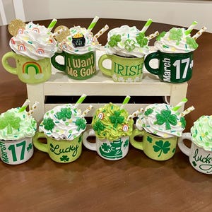May include: A collection of St. Patrick's Day themed mugs with whipped cream toppings, straws, and various decorations. The mugs are green and white, with text like "Lucky", "Irish", and "Kiss Me". Some mugs feature shamrocks and rainbows.