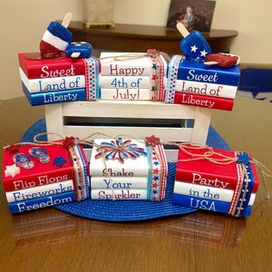 May include: A collection of six stacks of miniature wooden books painted in red, white, and blue. The books feature patriotic text including Sweet Land of Liberty, Happy 4th of July, Flip Flops, Fireworks, Freedom, Shake Your Sparkler, and Party in the USA.