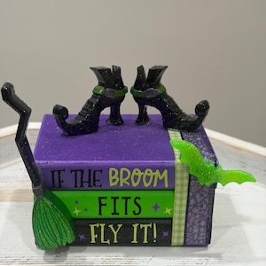 May include: A decorative stack of three books in purple, green, and black with the text IF THE BROOM FITS FLY IT. The stack is topped with two black glittery pointed boots and features a green glittery bat and a black broomstick attached.