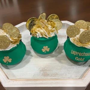 May include: Three green pot-shaped decorations with white frosting, gold coins, and glitter. Each pot has a shamrock design. One pot has the text "Leprechaun's Gold". The decorations are on a white wooden tray.