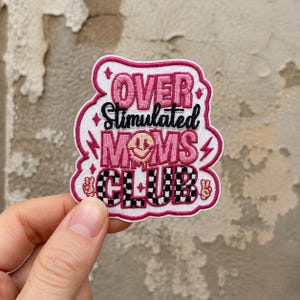 May include: An embroidered patch with a white background and pink border. It features the text OVER in pink, Stimulated in black, MOMS in pink with a smiley face, and CLUB in a black and white checkerboard pattern, accented by small stars and peace signs.