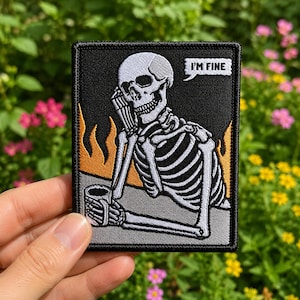 May include: A rectangular embroidered patch featuring a white skeleton sitting at a table with a mug, set against a black background with orange flames. A white speech bubble above the skeleton contains the black text I'M FINE.