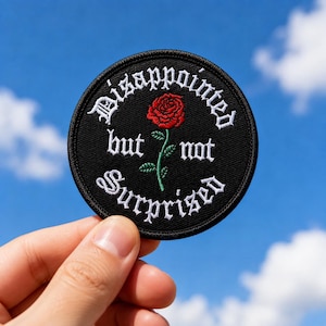 May include: A circular black embroidered patch featuring a red rose with a green stem in the center. The white text reads Disappointed but not Surprised in a gothic font style, arranged in a circular pattern around the central rose design.