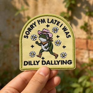 May include: A pale yellow embroidered patch with a green border featuring a frog wearing a pink cowboy hat, a brown neckerchief, and brown boots. The text reads SORRY I'M LATE I WAS DILLY DALLYING, surrounded by white daisy flowers and small black stars.