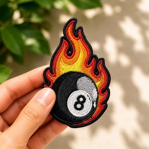 May include: An embroidered patch featuring a black billiard ball with a white circular center containing the number eight, surrounded by stylized flames in shades of red, orange, and yellow with a thick black border outline.