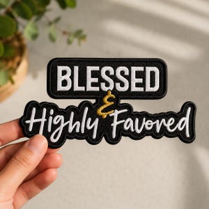 May include: A black embroidered patch featuring the white text BLESSED in a block font and the white script text Highly Favored, connected by a gold ampersand symbol. The patch has a black border and a distinct shape following the lettering.