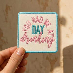 May include: A square embroidered patch with a cream-colored background and a light blue border. The text reads YOU HAD ME AT DAY drinking in a combination of pink block letters, pink cursive script, and a central teal block letter word DAY.