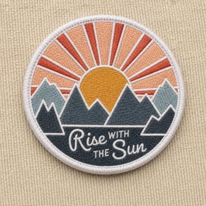 May include: A circular embroidered patch featuring a yellow sun rising over dark blue mountain peaks. The sky is composed of radiating peach and orange stripes. The text "Rise WITH THE Sun" is stitched in white across the lower dark blue section.