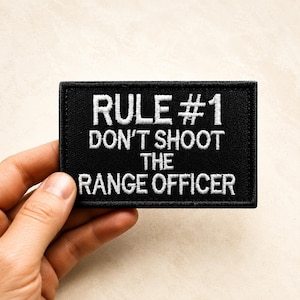 May include: A rectangular black embroidered patch with a stitched border. The white text reads: RULE #1 DON'T SHOOT THE RANGE OFFICER. The lettering is in a bold, sans-serif font, centered on the black fabric background of the patch.