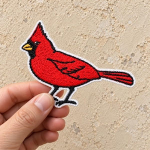 May include: An embroidered patch shaped like a northern cardinal bird. The bird features vibrant red thread, a black face mask around a yellow beak, and black stitched details on the wings and tail, all outlined with a white border.