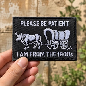 May include: A rectangular black embroidered patch featuring white stitching of an ox pulling a covered wagon. The text reads PLEASE BE PATIENT at the top and I AM FROM THE 1900s at the bottom. The patch has a black border and a textured fabric surface.