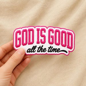 May include: A rectangular embroidered patch with a white background and a scalloped border. The text GOD IS GOOD is stitched in bold, bright pink block letters, while the phrase all the time is stitched in a black cursive script font.