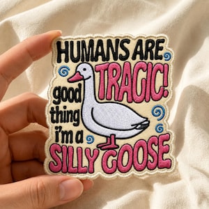 May include: A rectangular embroidered patch with a scalloped edge on a cream background. It features a white goose with a pink beak and feet, surrounded by black and pink text that reads: HUMANS ARE TRAGIC! good thing i'm a SILLY GOOSE.