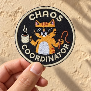 May include: A circular embroidered patch with a black background and a cream-colored border. It features an orange tabby cat wearing black sunglasses, holding a steaming mug of coffee in one hand and a brown whip in the other, with the text CHAOS COORDINATOR.