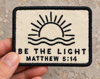 May include: A rectangular embroidered patch with a cream-coloured background and a thick black border. The design features a black embroidered sun rising over wavy lines, with the text BE THE LIGHT and MATTHEW 5:14 stitched in black below.