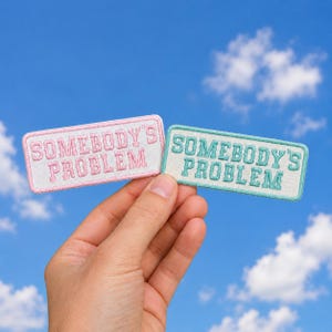 May include: Two rectangular embroidered patches with white backgrounds and stitched borders. One patch features pink text reading SOMEBODY'S PROBLEM, while the other features teal text reading SOMEBODY'S PROBLEM in a block letter style.