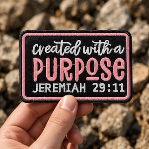 May include: A rectangular embroidered patch with a black background and a pink border. The text reads "created with a" in white script, "PURPOSE" in large pink block letters, and "JEREMIAH 29:11" in white block letters at the bottom.
