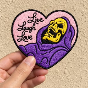 May include: A heart-shaped embroidered patch featuring a yellow skull character wearing a purple hood. The background of the patch is light pink with the black embroidered text "Live Laugh Love" written in a cursive script on the left side.