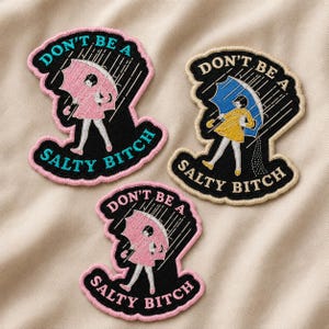 May include: Three embroidered patches featuring a figure holding an umbrella in the rain. Two patches show a pink umbrella and dress with pink borders, while one shows a blue umbrella and yellow dress with a cream border. All include the text DON'T BE A SALTY BITCH.