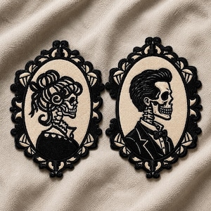 May include: Two oval embroidered patches featuring black and cream thread. One patch depicts a skeleton with an updo hairstyle, the other a skeleton in a tuxedo. Both are enclosed in ornate, decorative black frames with intricate scrollwork details.