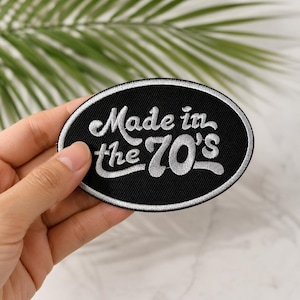 May include: An oval-shaped embroidered patch with a black background and a silver border. The text "Made in the 70's" is stitched in silver thread across the center in a stylized, cursive font. The patch features a clean, finished edge.