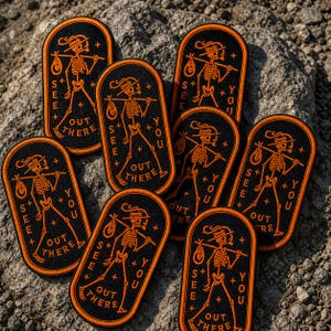 May include: Seven oval-shaped black embroidered patches with orange borders. Each patch features a skeleton wearing a hat, carrying a bindle on a stick, and the text "SEE YOU OUT THERE" with small plus signs arranged around the central figure.