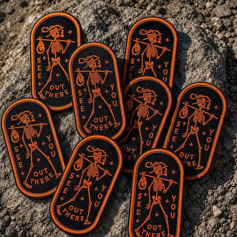 May include: Seven oval-shaped black embroidered patches with orange borders. Each patch features a skeleton wearing a hat, carrying a bindle on a stick, and the text "SEE YOU OUT THERE" with small plus signs arranged around the central figure.