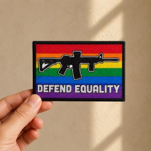 May include: A rectangular embroidered patch featuring a horizontal rainbow flag background with a black silhouette of a rifle centered in the middle. The text DEFEND EQUALITY is stitched in white capital letters along the bottom edge.
