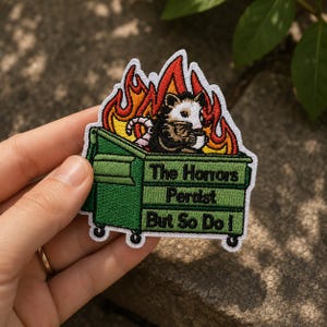 May include: An embroidered patch featuring a green dumpster with a white and black opossum sitting inside amidst orange and red flames. The text on the dumpster reads, The Honors Persist But So Do I, in black thread against the green background.