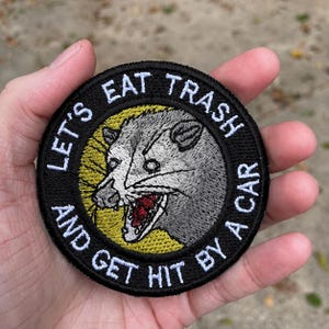 May include: A circular embroidered patch featuring a gray opossum with an open mouth showing teeth against a yellow background. The black border contains the white embroidered text: LET'S EAT TRASH AND GET HIT BY A CAR.