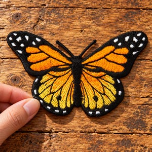May include: An embroidered butterfly patch featuring vibrant orange and yellow wings with black borders and white spots. The design includes a black central body and two antennae, showcasing detailed stitching throughout the entire insect shape.
