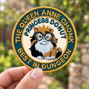 May include: A circular embroidered patch featuring a calico cat wearing a gold crown and teal glasses. The text reads THE QUEEN ANNE OHONK PRINCESS DONUT BEST IN DUNGEON. The patch has a cream center, a dark teal border, and a gold outer edge.