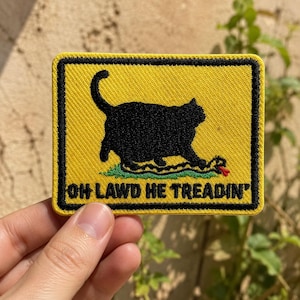 May include: A rectangular embroidered patch with a yellow background and a black border. It features a black silhouette of a cat standing on a snake, with the text "OH LAWD HE TREADIN'" printed in black capital letters at the bottom of the patch.